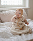 A baby in a beige outfit and knitted hat sits on white bedding, smiling and holding a FRIGG Andersen Fairytale Natural Rubber Pacifier 2-Pack by FRIGG, with soft natural light streaming in from the window behind.