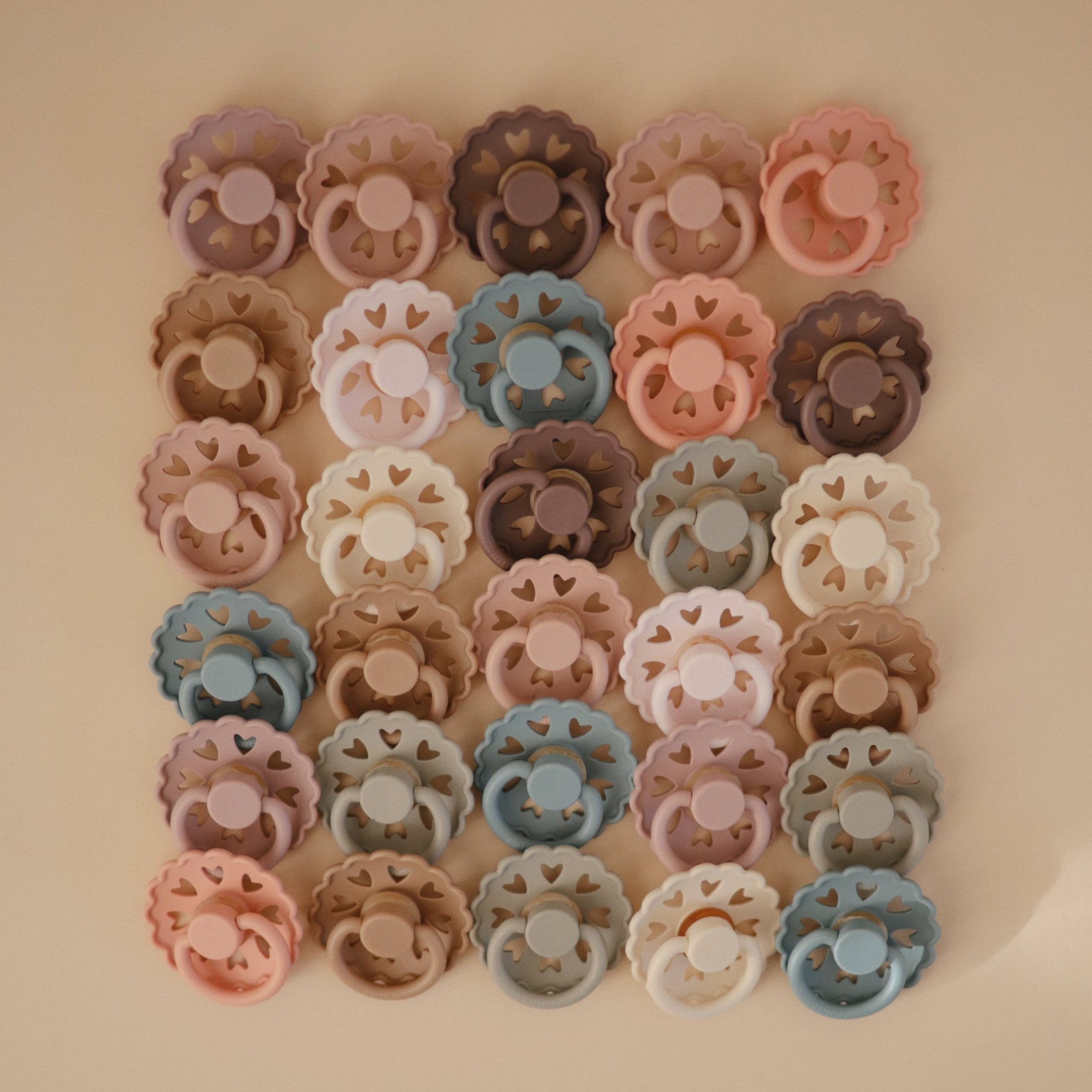 Lifestyle image of Frigg Andersen pacifiers laid out in a square grid with cream, blush, croissant, grey, blue, and purple pacifiers. 