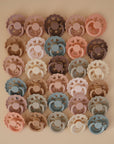 Lifestyle image of Frigg Andersen pacifiers laid out in a square grid with cream, blush, croissant, grey, blue, and purple pacifiers. 