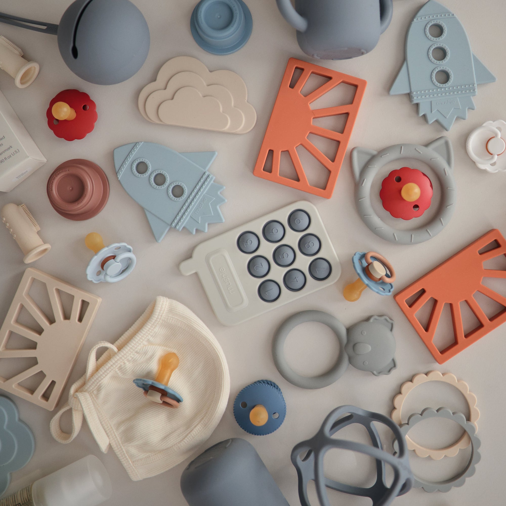 A flat lay of pastel-colored baby items neatly arranged on a light surface, featuring the mushie Sun Teether alongside pacifiers, rings, a cloud-shaped piece, and a small fabric pouch.