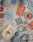 A flat lay of pastel-colored baby items neatly arranged on a light surface, featuring the mushie Sun Teether alongside pacifiers, rings, a cloud-shaped piece, and a small fabric pouch.