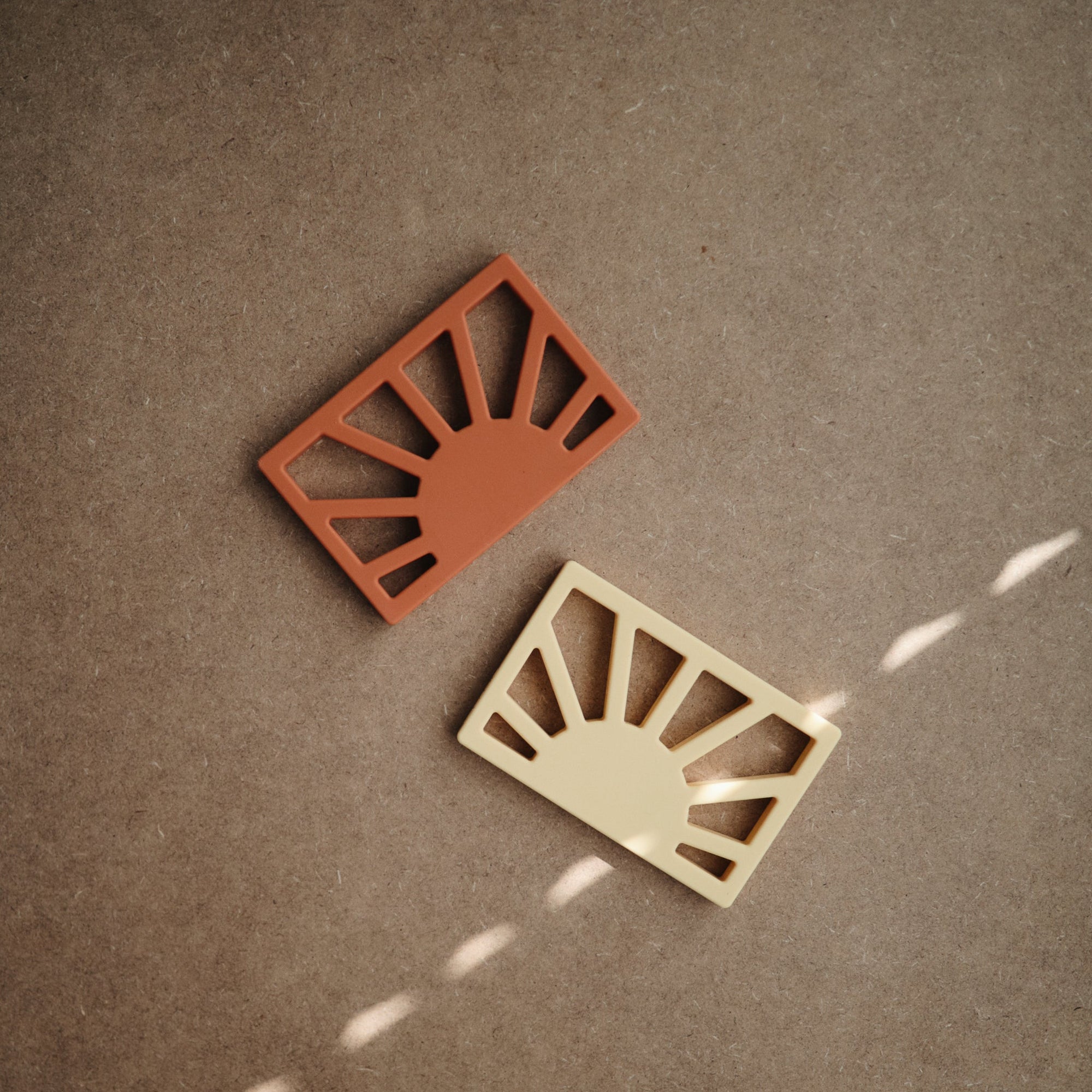Two mushie Sun Teethers in terracotta and beige, both featuring sunburst designs, rest on a brown textured surface with light streaks. These non-toxic teethers provide style and safety for your little one.