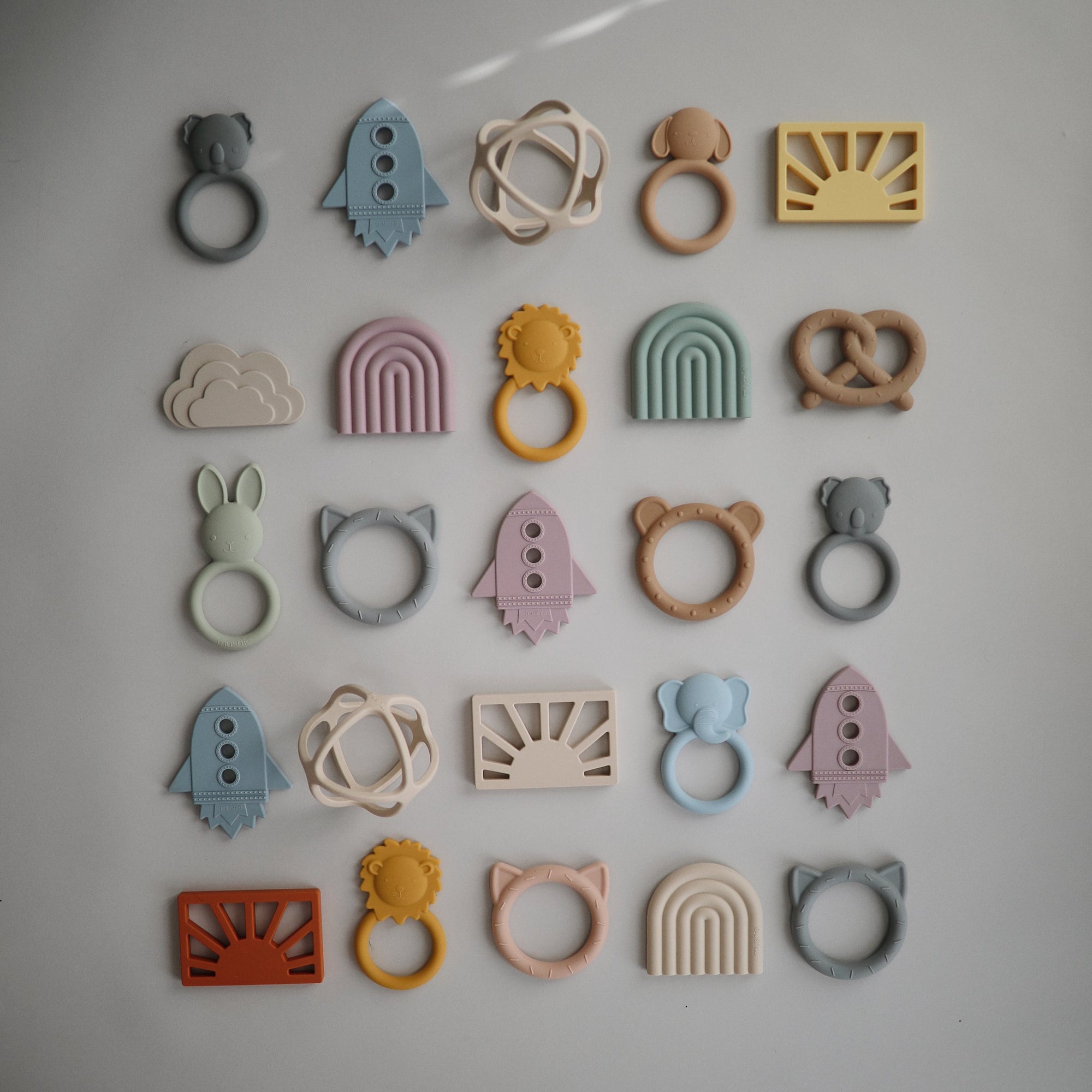 A neatly arranged grid of mushie Sun Teether and other colorful, non-toxic silicone baby teethers in fun shapes—rockets, rainbows, clouds, animals, pretzels—displayed on a neutral background.