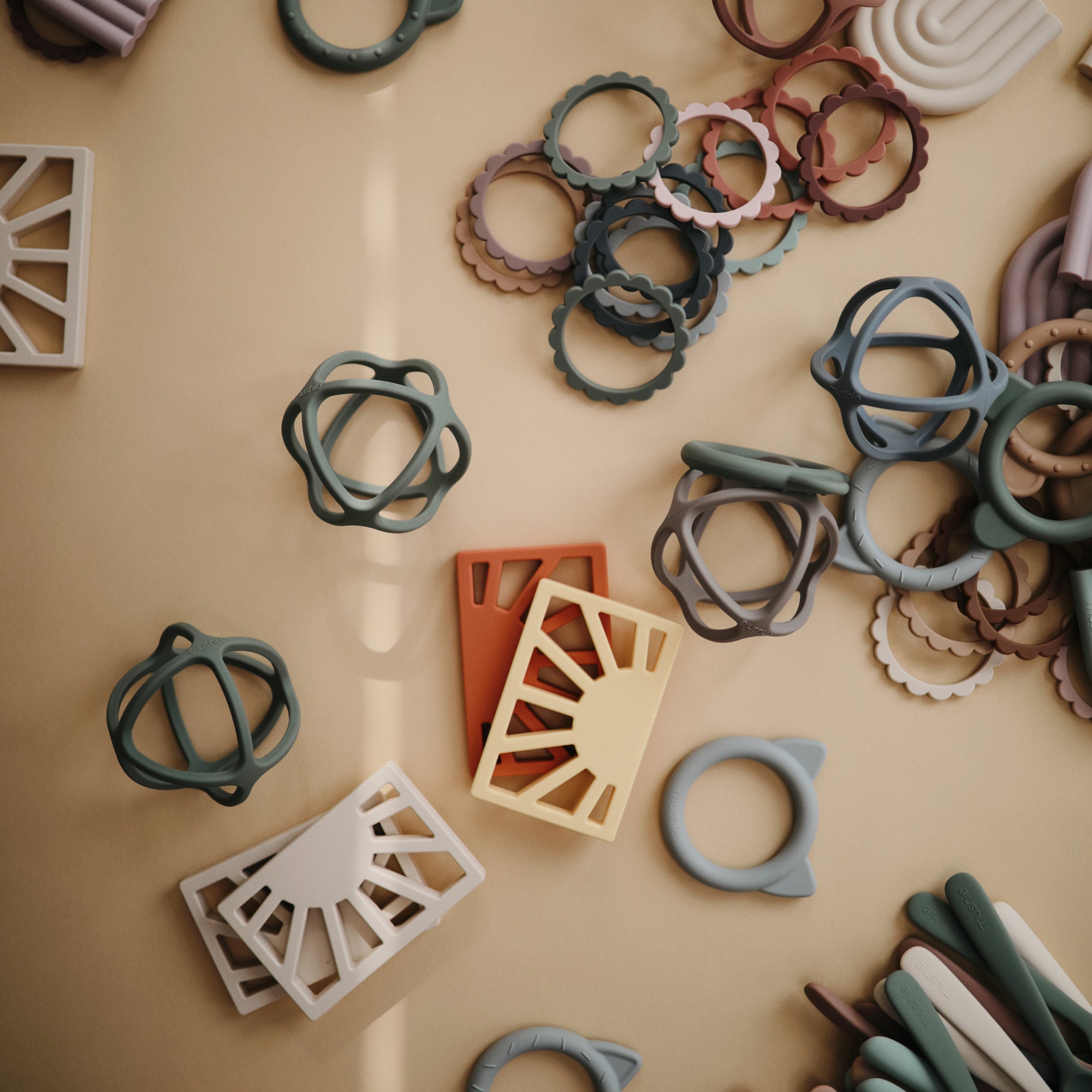 The mushie Sun Teether and other assorted silicone baby toys in soft, muted colors are scattered on a beige surface, featuring non-toxic materials and safe rounded or geometric designs for playful teething and stacking.
