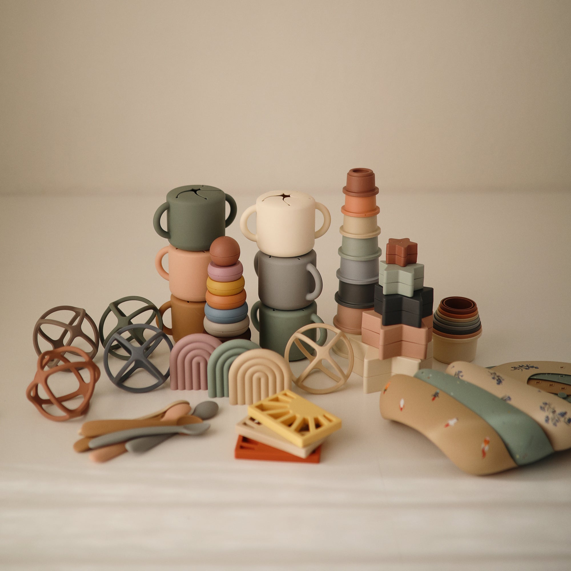 The mushie Sun Teether, a pastel-colored silicone baby toy, is neatly displayed on a light surface alongside stacking cups, building blocks, utensils, rainbow arches, and soft blocks.