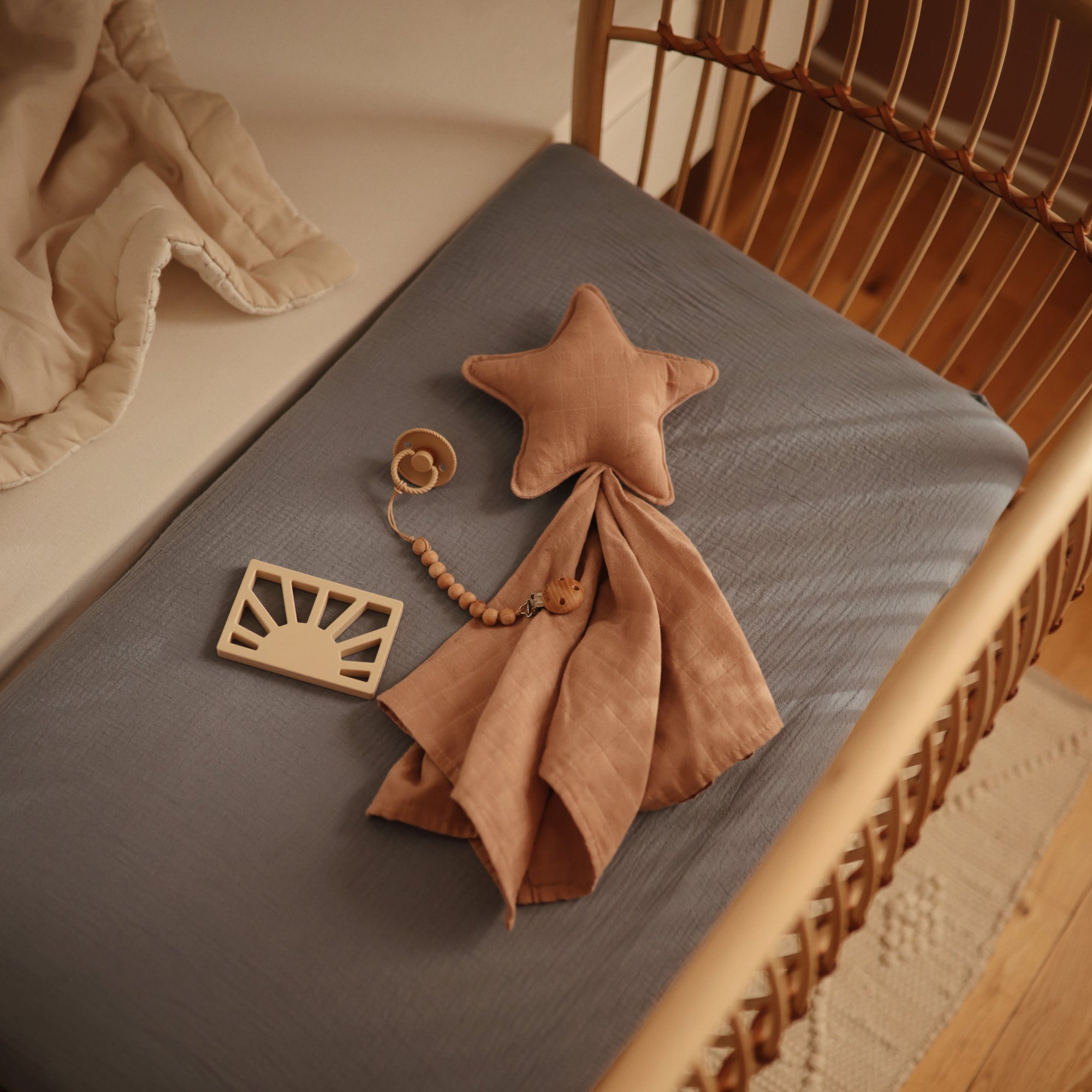 A cozy nursery featuring a rattan crib, blue mattress, beige blanket, star-shaped soft toy with pink cloth, wooden pacifier clip, and the mushie Sun Teether along with a silicone baby teether on the mattress.